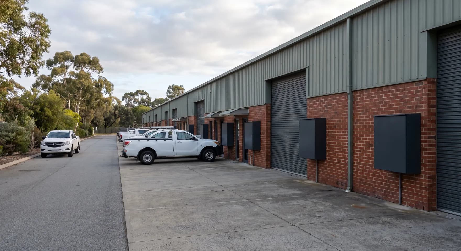 Wall mounted chargers beside the roller doors of a light industrial unit row in O'Connor, with utes parked on the shared apron